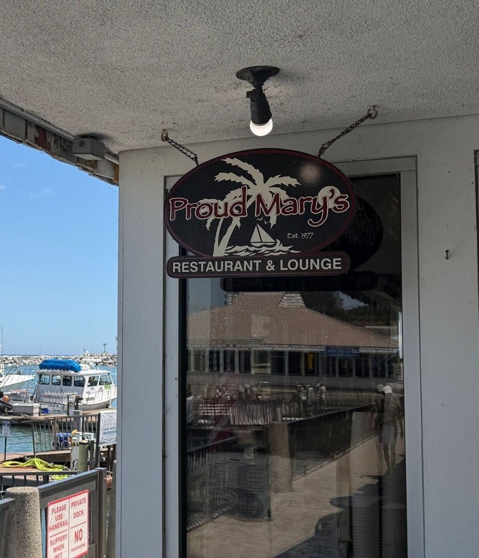 Proud Mary’s Restaurant & Lounge oval sign Est. 1977 at Dana Point Harbor with boats beyond